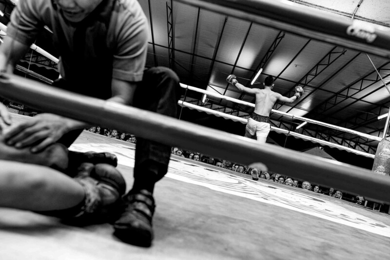 Muay Thai ringinde yerde yatan bir dövüşçüye eğilen görevli ve arka planda kollarını açmış ayakta duran sporcu