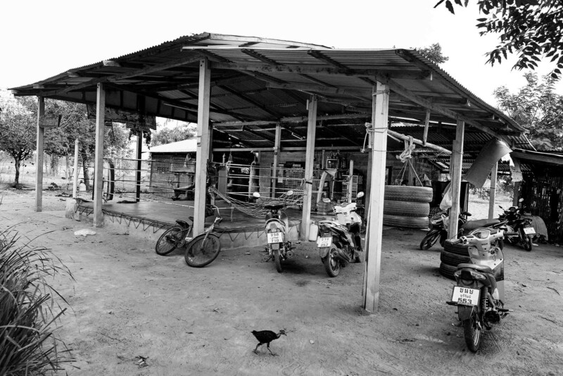 Tayland kırsalında açık kenarlı, sac çatılı mütevazı bir Muay Thai antrenman alanı ve önünde park edilmiş motosikletler