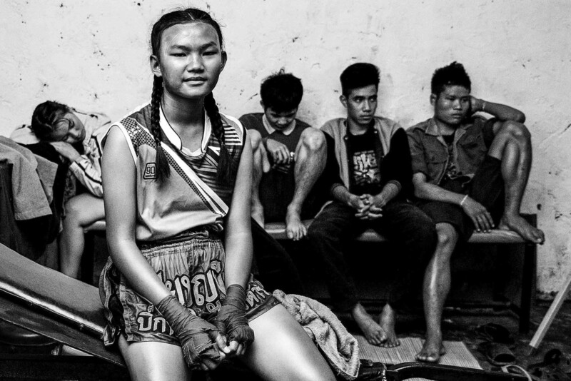 Ellerinde bandaj bulunan genç bir Muay Thai sporcusu, dinlenen diğer sporcuların önünde oturuyor