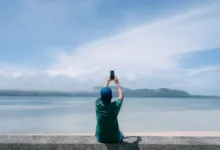 Seyahat fotoğrafçılığı için hangi mobil aksesuarlar iyidir? 29 person sits on wall in front of water while holiding up their phone