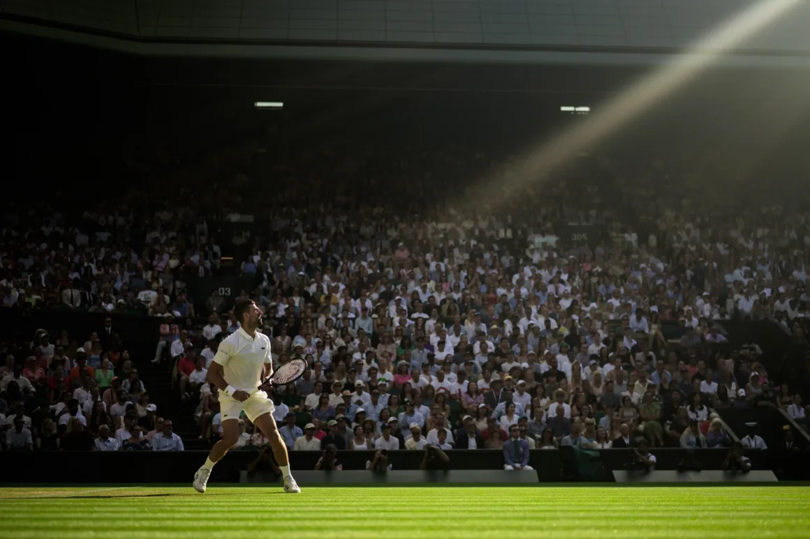 a beam of sunlight illuminates a tennis player on a grass court with stands filled behind him