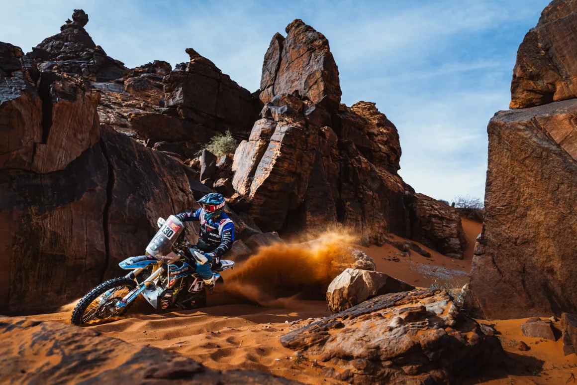 a person on a blue dirt bike blows up sand while riding through a rocky gap