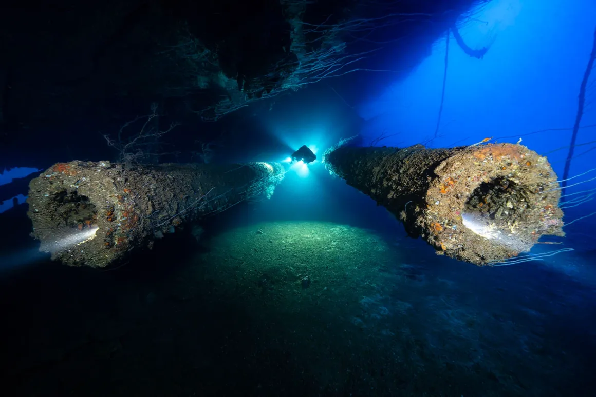 A scuba diver explores the seafloor near two large, encrusted pipes or cannons extending from a sunken shipwreck, illuminated by the diver's bright flashlight against deep blue water.