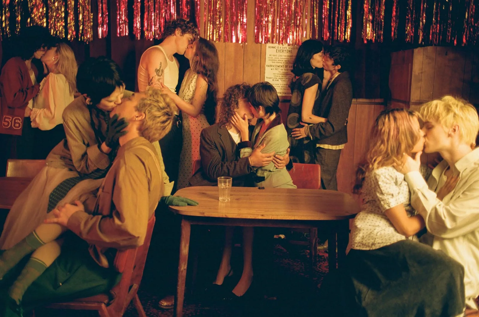 Loş ışıklı, şenlikli bir odada genç çiftlerden oluşan bir grup tutkuyla öpüşüyor; duvarda kırmızı simli süsler asılı, etraftaki insanlar ahşap bir masanın çevresinde oturuyor ya da ayakta duruyor; samimi ve canlı bir atmosfer oluşuyor.