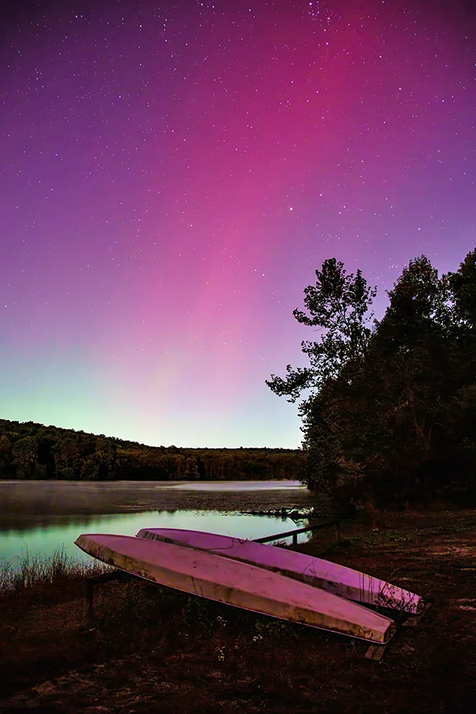 A vibrant night sky glows pink and purple above a calm lake, with two upside-down boats resting on the grassy shore beside trees and distant hills.