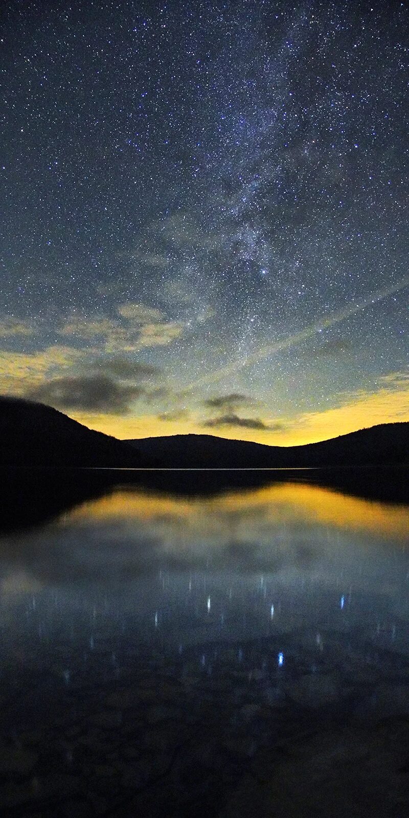 A star-filled night sky is reflected on a tranquil lake, with distant hills silhouetted against a faint golden glow on the horizon. The Milky Way is visible among the stars above.