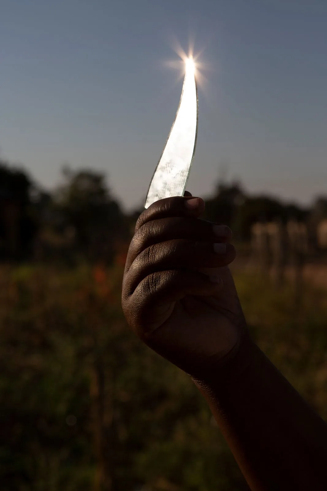 Bir el, kavisli bir cam parçasını kaldırıp tutuyor; güneşi yakalayan camın ucunda parlak, yıldız benzeri bir ışıltı oluşuyor. Arka planda dış mekân, bulanık halde görünüyor.