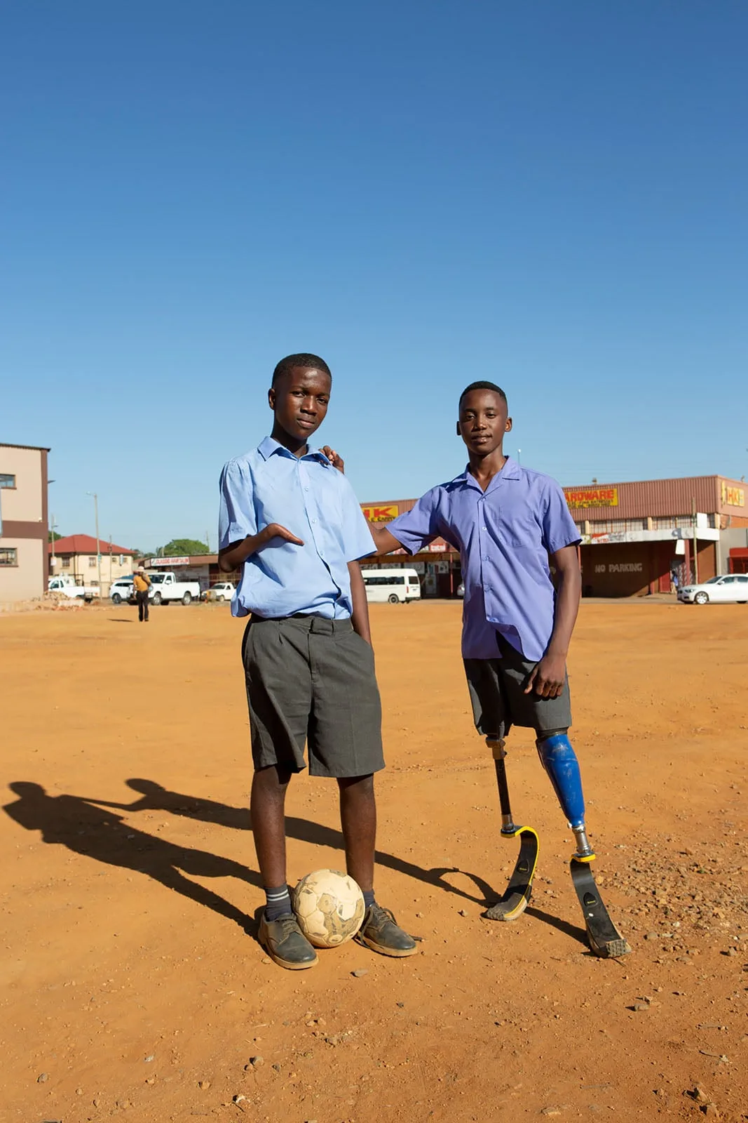 Tozlu bir sahada, bir futbol topunun yanında iki çocuk duruyor. İkisi de okul üniforması giyiyor; birinin protez kolu, diğerinin ise iki protez bacağı var. Arka planda binalar ve arabalar, berrak mavi bir gökyüzü altında görünüyor.