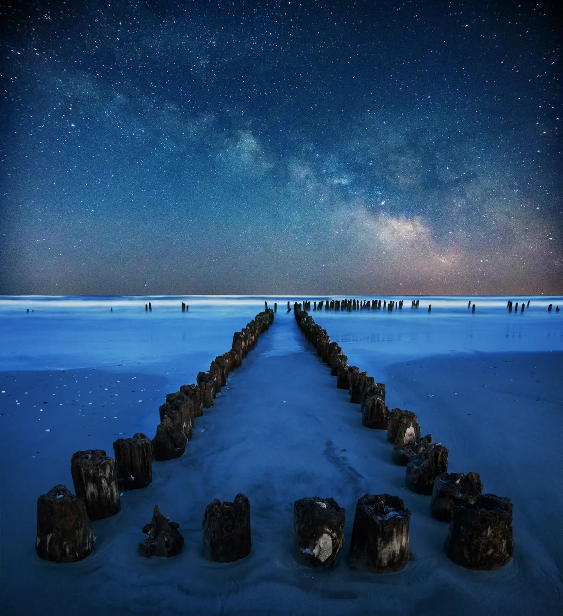Old wooden posts form two parallel lines on a sandy beach at night, leading toward the ocean. The sky is filled with stars and the Milky Way, creating a serene and mystical atmosphere.