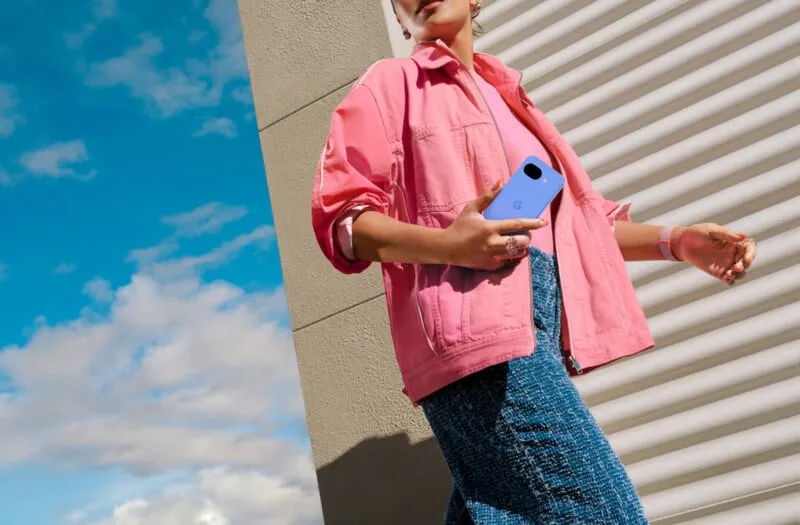 Pembe ceket ve mavi etek giyen bir kişi dış mekânda açık mavi bir akıllı telefon tutuyor. Arka planda dokulu bir duvar, metal bir kepenk ve parçalı bulutlu mavi gökyüzü görülüyor.