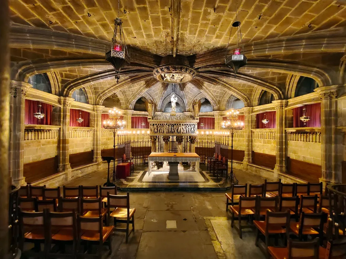 Vivo V70 İncelemesi: Daha İyi Yazılım, Daha İyi Tasarım ve Küçük Adımlar 13 Tonozlu tavanlara sahip taş bir şapel; süslü avizeler, ahşap sandalyeler, ortada bir altar, duvarlarda kırmızı perdeler ve altarın üzerinde bir heykel ile sıcak ışık altında detaylı bir iç mekân.