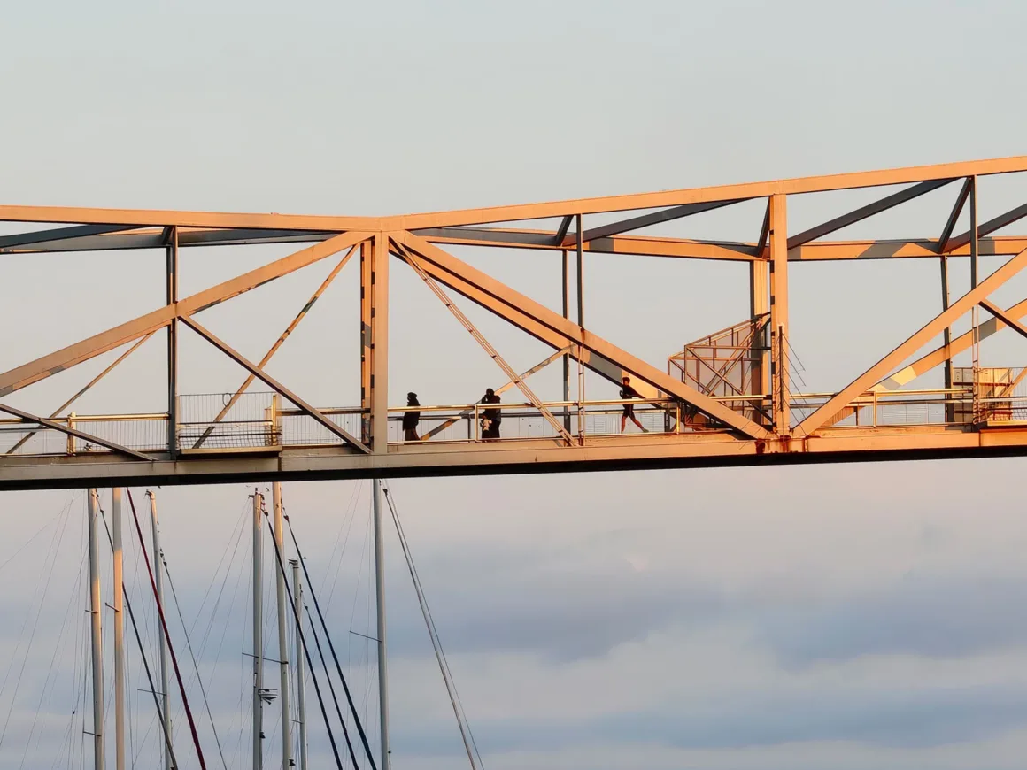 Vivo V70 İncelemesi: Daha İyi Yazılım, Daha İyi Tasarım ve Küçük Adımlar 9 Gün batımında, sıcak ışıkla aydınlanan yükseltilmiş bir yaya köprüsünden geçen insanlar; aşağıda yelkenli teknelerin direkleri bulutlu gökyüzüne karşı siluet oluşturuyor.