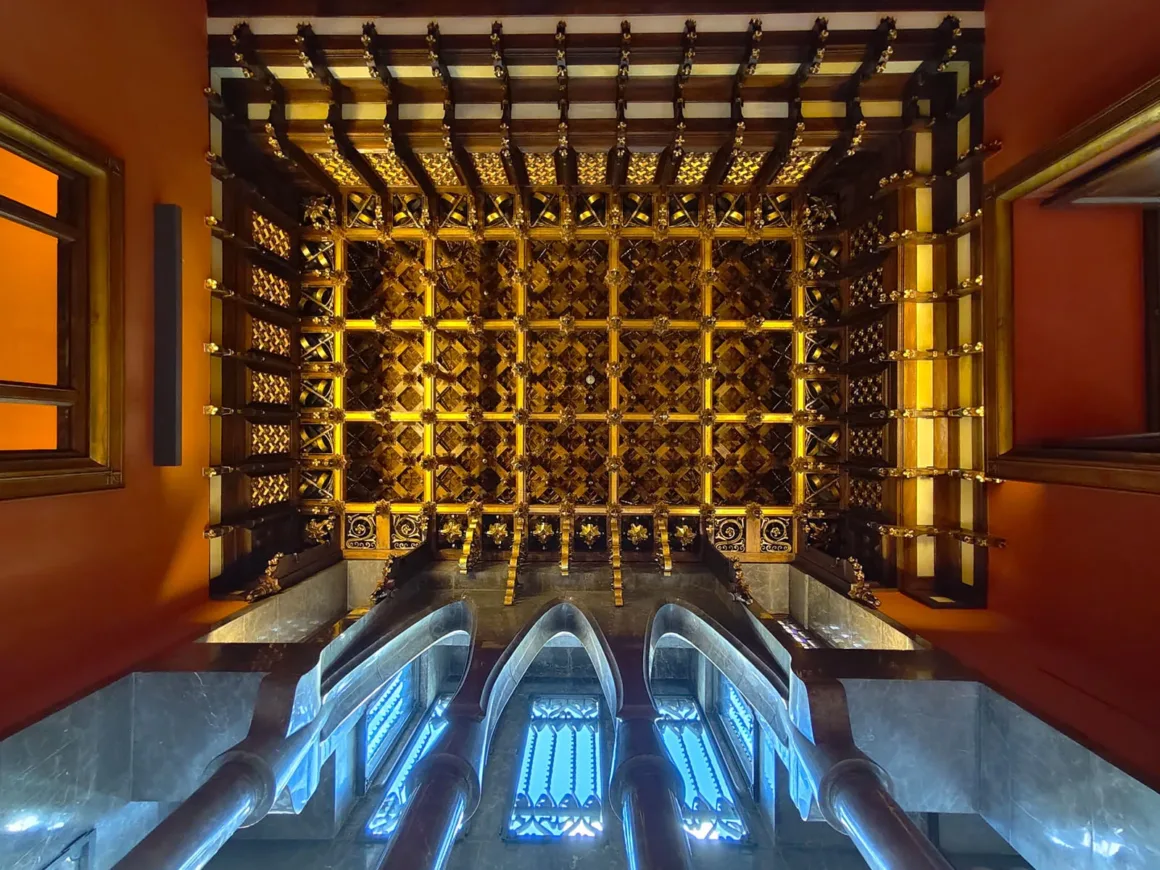 Vivo V70 İncelemesi: Daha İyi Yazılım, Daha İyi Tasarım ve Küçük Adımlar 11 Aşağıdan bakıldığında görülen, altın detaylı karmaşık bir tavan; kırmızı ve turuncu duvarlar, kemerli pencereler ve mavi tonlu mermer sütunlarla çevrili.