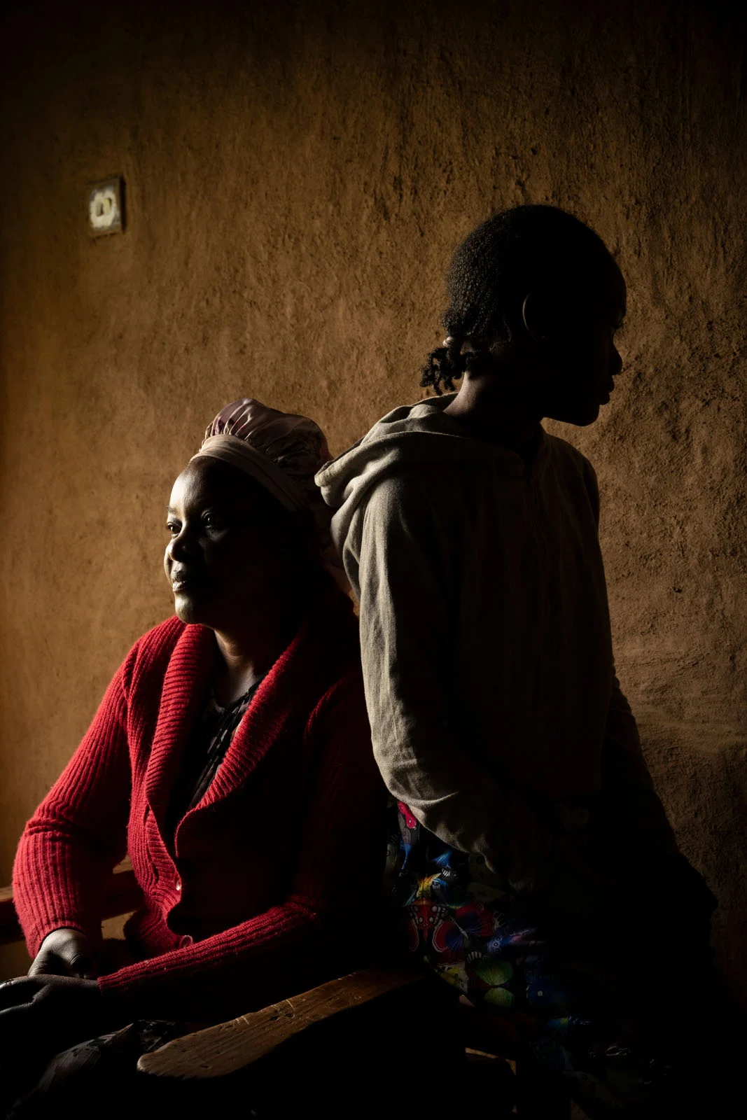 Kahverengi duvarlara sahip loş bir odada, kırmızı kazaklı bir kadın ve kapüşonlu bir kız yan yana. Kadın ışığa dönük, kız ise gölgede; ifadeleri sakin ve düşünceli.