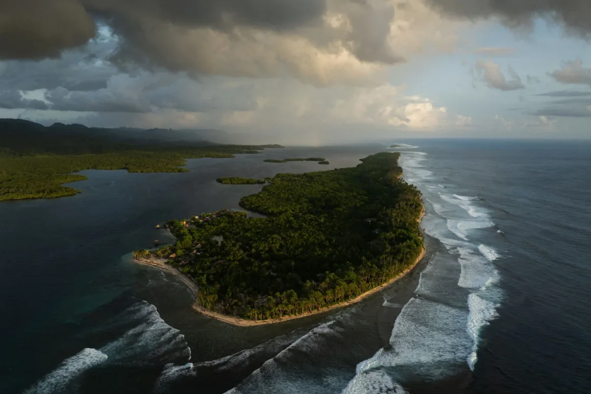 Yoğun ormanlarla kaplı, kıyı yerleşimleri bulunan ve okyanus dalgalarıyla çevrili yemyeşil bir yarımadanın havadan görünümü; dramatik bulutlar manzara üzerine gölgeler düşürüyor.