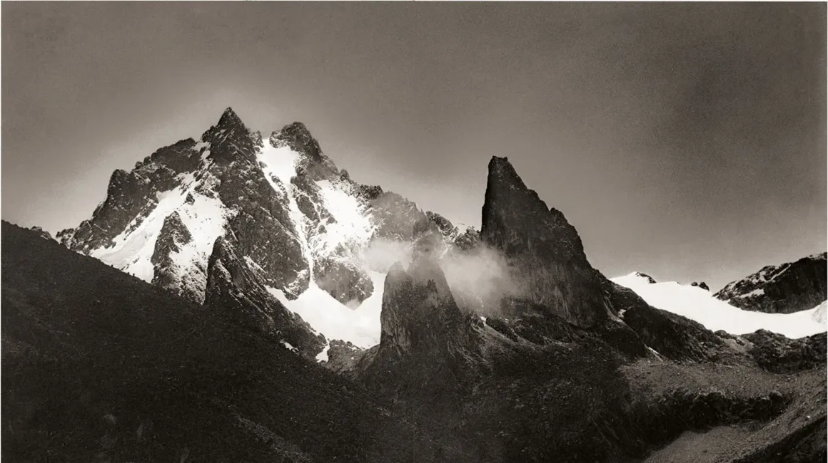 Karlarla kaplı, keskin ve engebeli dağ zirveleri bulutlu bir gökyüzüne doğru yükseliyor. Zirvelerin etrafında dolaşan sis ve bulutlar, dramatik manzarayı vurguluyor.