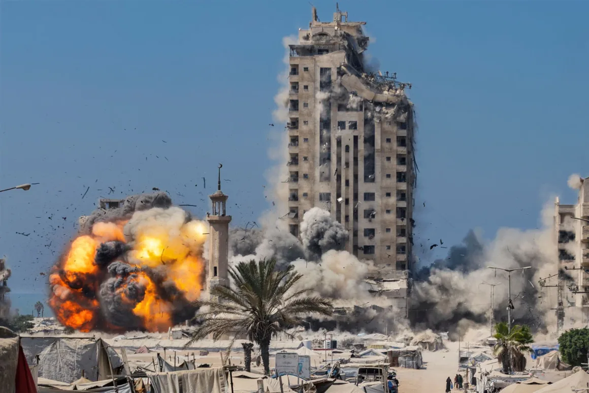 Bir yüksek binanın tabanında büyük bir patlama gerçekleşiyor; alevler, duman ve enkaz havaya savruluyor. Alt bölgede çadırlar, palmiye ağaçları ve elektrik hatları açık mavi gökyüzü altında görülüyor.