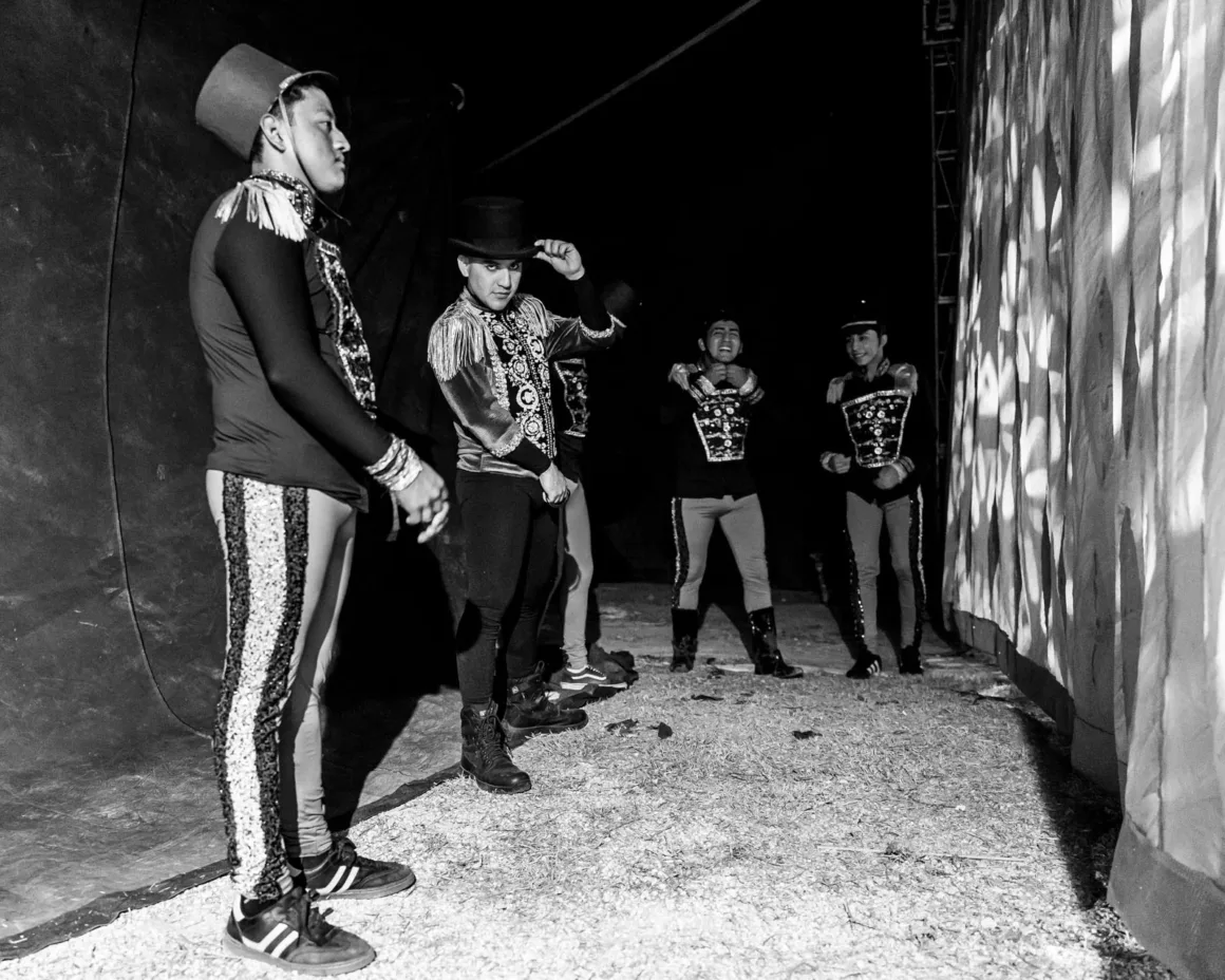 Fotoğrafın Acımasız Matematiğiyle Barışmak: Kontrol Bir Yanılsama mı? 2 Dekoratif kostümler ve silindir şapkalar giyen beş erkek sirk performansçısı çadırın arkasında bekliyor. Bazıları kıyafetlerini düzeltiyor, bazıları ise dramatik ışık ve gölgeler altında bekliyor.