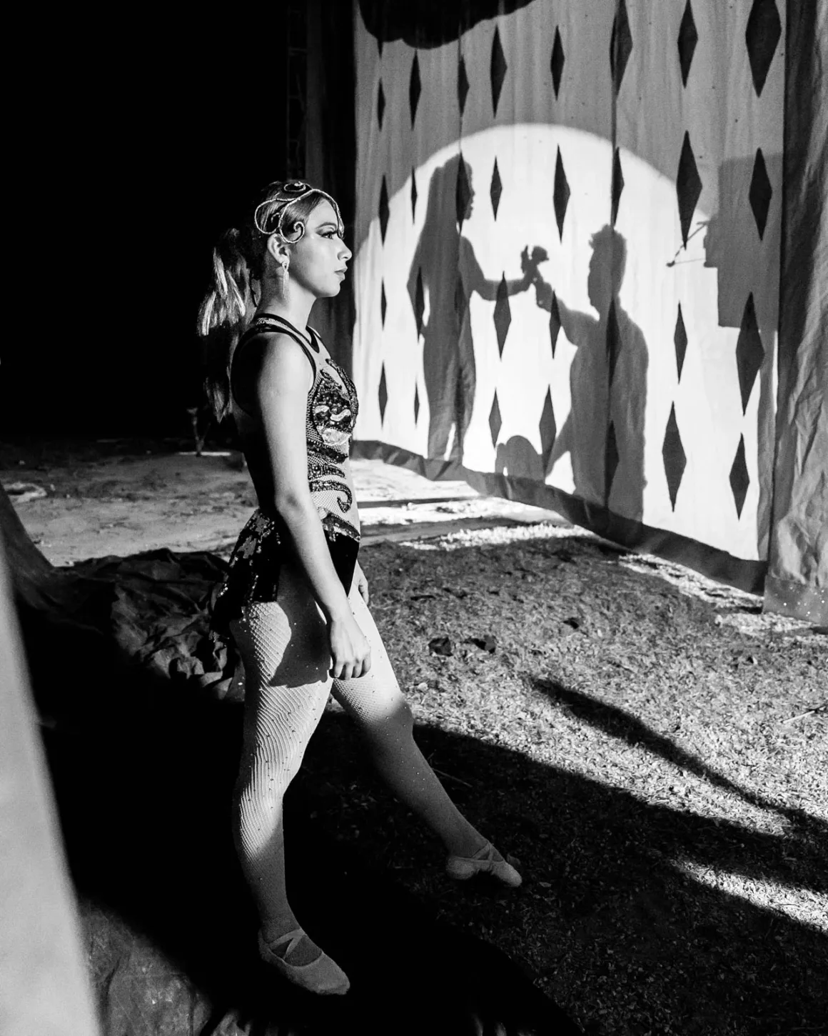 Fotoğrafın Acımasız Matematiğiyle Barışmak: Kontrol Bir Yanılsama mı? 5 Kostümlü bir sirk performansçısı kuliste duruyor ve elmas desenli perdelerle aydınlatılmış sahneye bakıyor. Spot ışığı arkasında güçlü bir gölge oluşturuyor. Siyah beyaz.