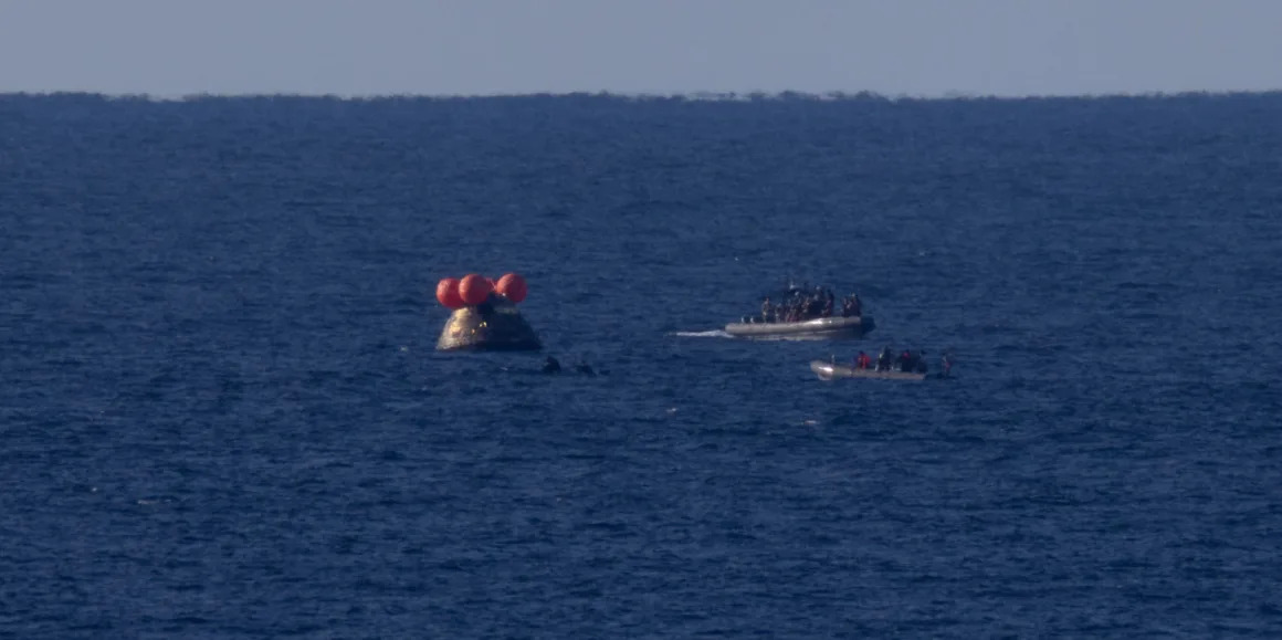 Turuncu şamandıralarla desteklenen kapsül denizde yüzerken, içinde insanlar bulunan tekneler yaklaşır; açık bir gökyüzü ve ufuk çizgisi.