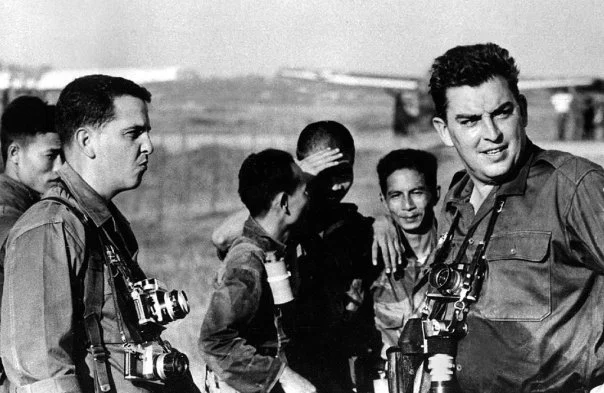 Bir grup foto muhabiri ve yerel adam açık havada duruyor; bazılarının boynunda kameralar asılı. İçlerinden biri gözlerini güneşten korurken diğerleri farklı yönlere bakıyor; hepsi dikkatli ve tetikte görünüyor.