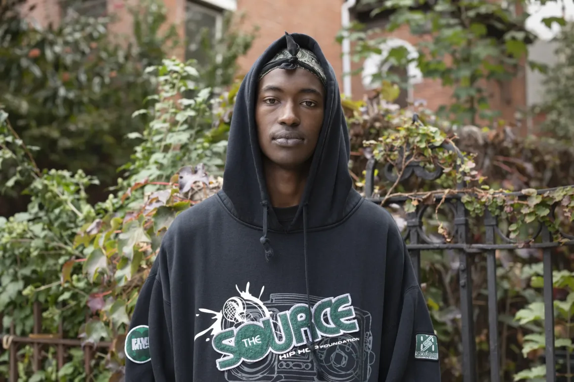Üzerinde 'The Source Hip Hop Music Awards' grafiği bulunan siyah bir hoodie giyen genç bir kişi, yapraklı çalılar ve tuğla bir bina önünde dış mekânda duruyor ve doğrudan kameraya bakıyor.