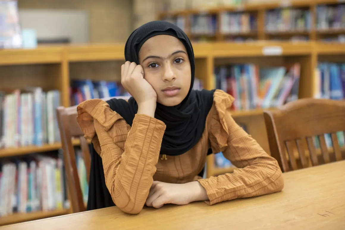 Siyah başörtüsü ve kahverengi bir üst giyen genç bir kız, bir kütüphanede masada oturuyor. Başını eline yaslamış şekilde düşünceli bir ifadeyle kameraya bakıyor. Arka planda kitap rafları görülüyor.