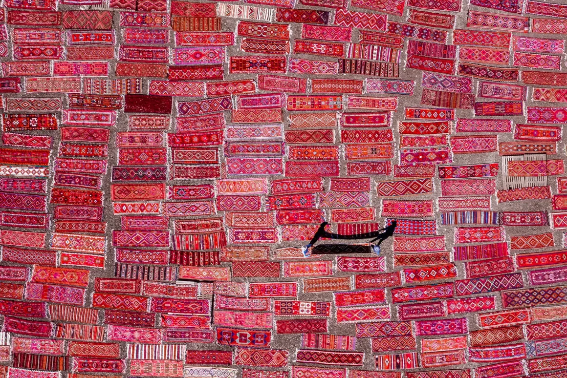 Duvarı kaplayan canlı kırmızı ve pembe tonlarda, desenli halıların oluşturduğu büyük bir düzenleme; ortada tek bir siyah minderli oturma alanı bulunuyor. Her halı geleneksel motiflerle süslenmiş.