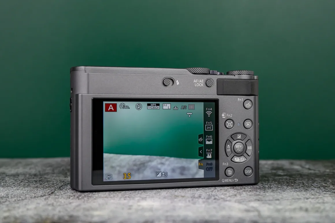 Panasonic Lumix ZS300 İnceleme: Eski Model Daha İyiydi? 10 Dokulu bir yüzey üzerinde duran bir dijital kamera, arka yüzü izleyiciye dönük şekilde gösteriliyor. Ekranda kamera ayarları ve çeşitli kontrol düğmeleri görülüyor. Arka plan koyu yeşil.