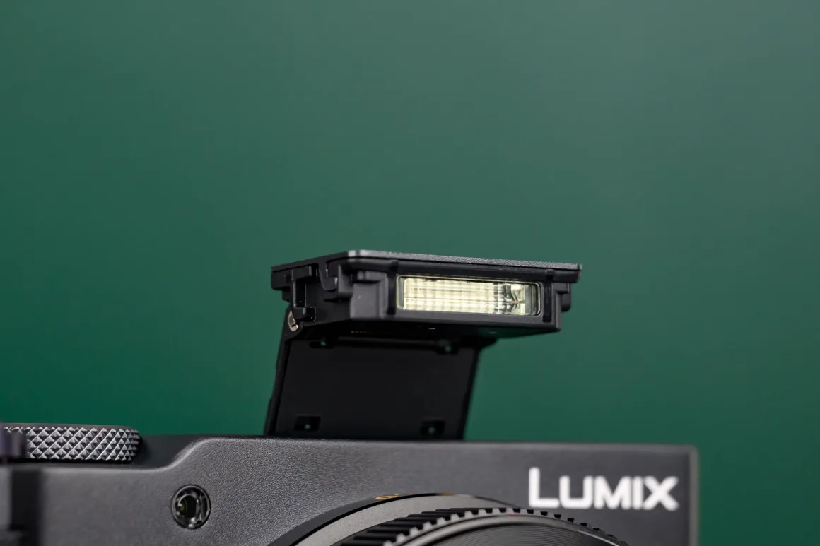 Panasonic Lumix ZS300 İnceleme: Eski Model Daha İyiydi? 9 Sade yeşil bir arka plan önünde dahili flaşı açık durumda olan siyah bir Lumix kameranın yakın planı.
