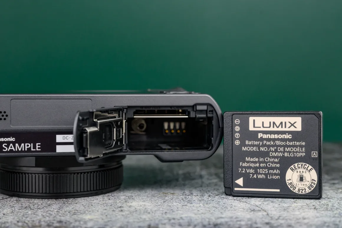 Panasonic Lumix ZS300 İnceleme: Eski Model Daha İyiydi? 5 Pil bölmesi açık bir kameranın yakın planı; yanında gri yüzey üzerinde dik duran Panasonic Lumix DMW-BLG10PP şarj edilebilir lityum iyon pil bulunuyor.
