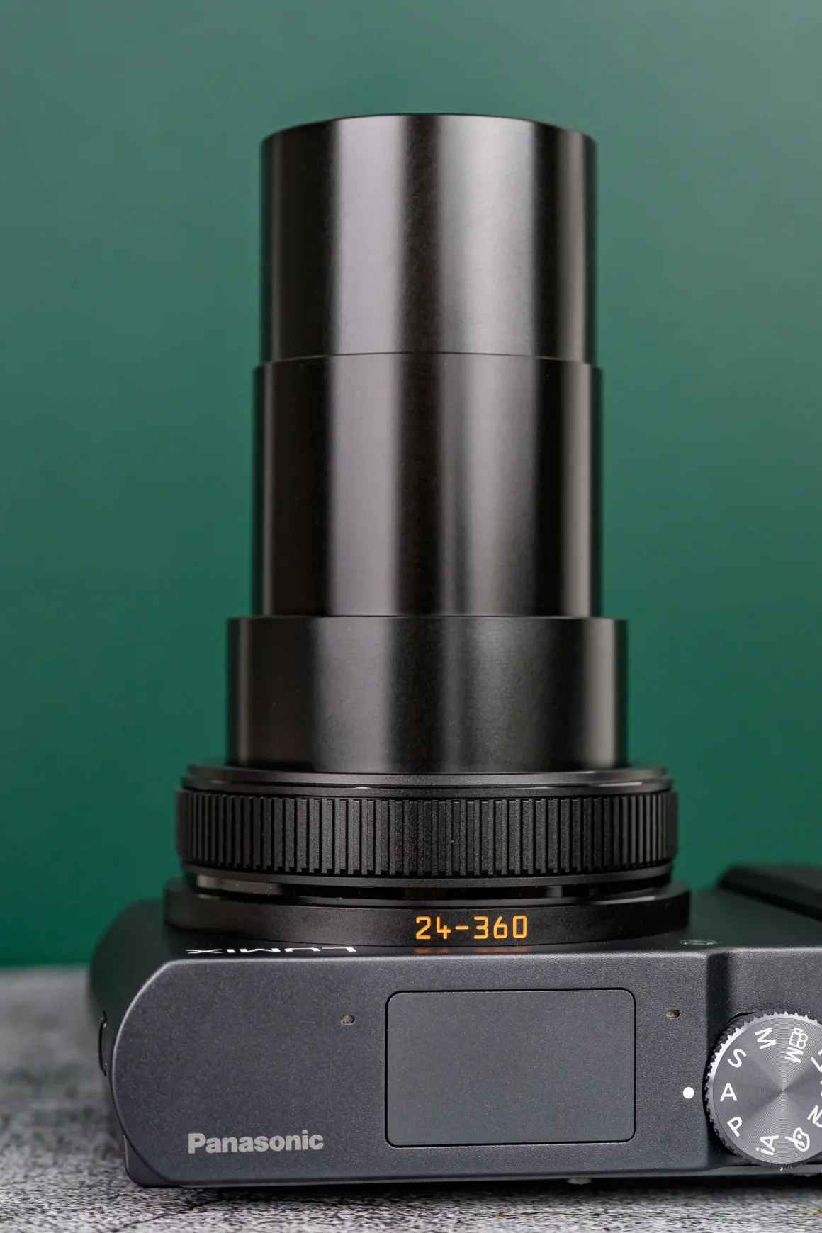 Panasonic Lumix ZS300 İnceleme: Eski Model Daha İyiydi? 7 Siyah bir Panasonic dijital kameranın yakın planı; zoom lensi tamamen uzatılmış ve lens halkasında "24-360" yazısı görülüyor. Arka plan yeşil.