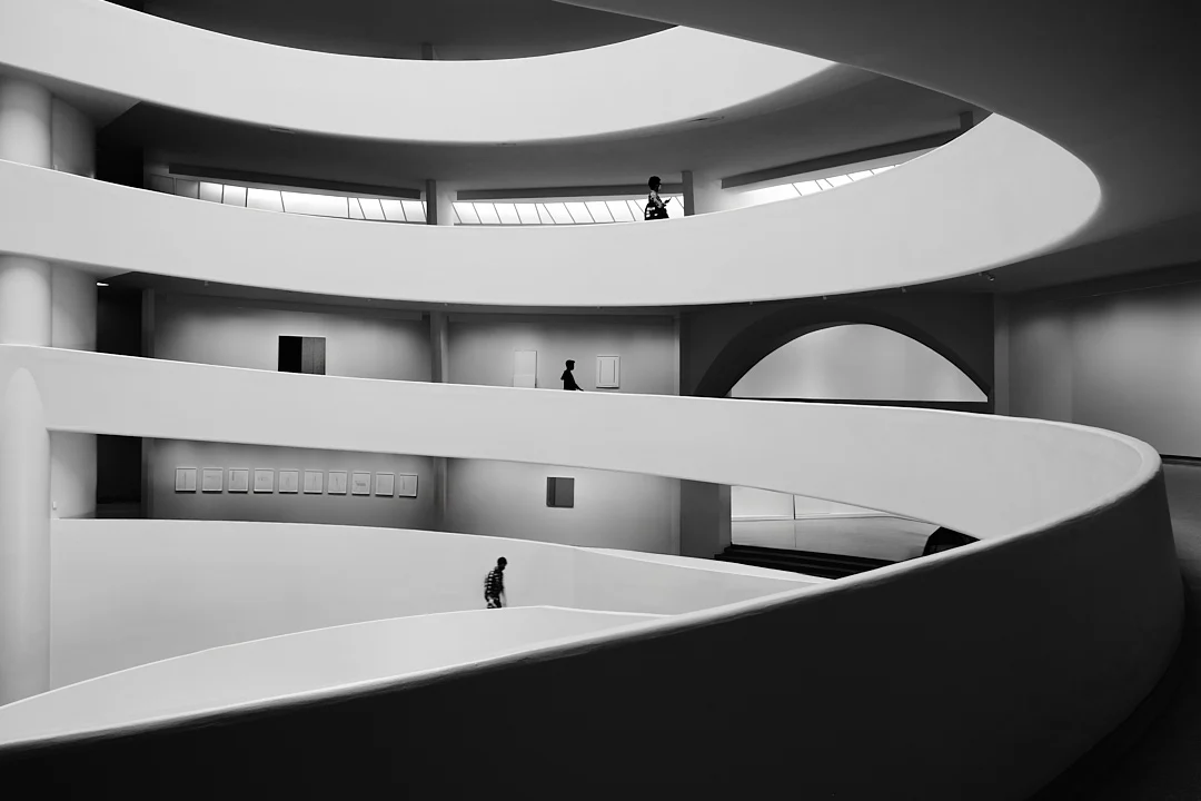 Kıvrımlı rampalara, geometrik formlara ve farklı katlarda yürüyen üç kişiye sahip modern, dairesel bir iç mekânın siyah beyaz fotoğrafı; ölçek ve mimari derinlik hissi yaratıyor.