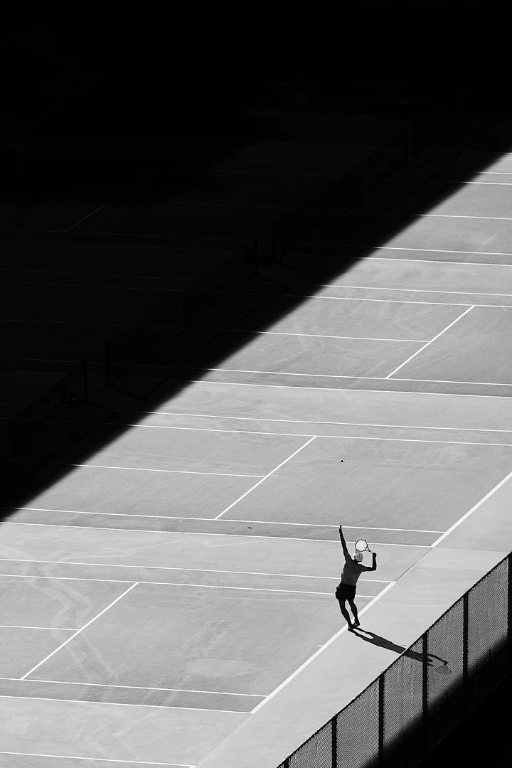 Güneş ışığında servis atmaya hazırlanan bir tenisçi, uzun bir gölge oluşturuyor. Çevredeki kortlar gölgede kalırken ışık ve karanlık arasında güçlü bir kontrast oluşuyor.