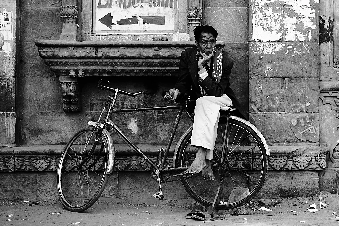 Eski, dokulu bir taş duvara yaslanmış bisikletin üzerinde oturan bir adam. Dirseğini dizine dayamış, yüzünü eline yaslamış ve düşünceli bir şekilde bakıyor. Siyah beyaz sahnede yıpranmış yüzeyler ve tabelalar dikkat çekiyor.