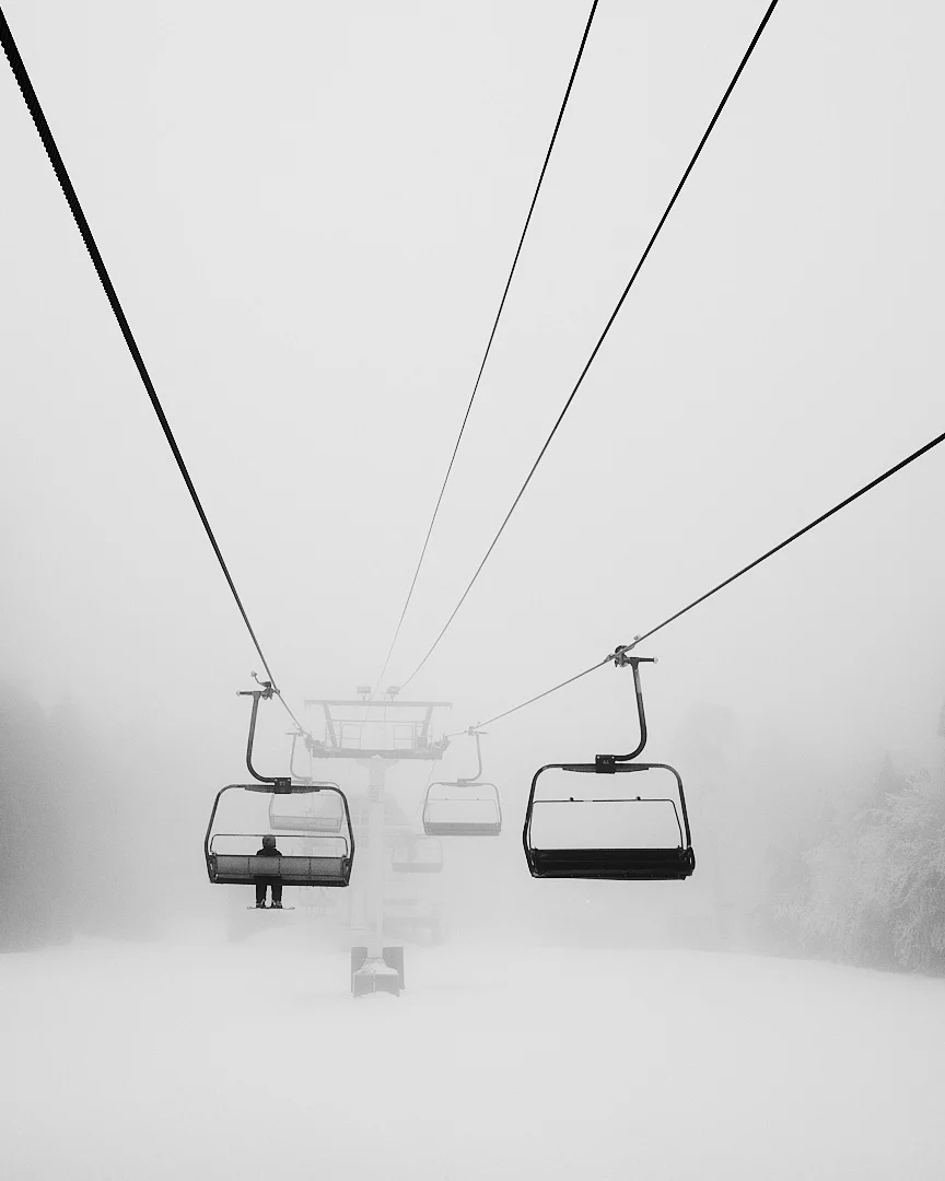 Karla kaplı bir manzaranın üzerinde yoğun sisin içinden yükselen neredeyse boş bir telesiyej; sadece bir sandalyede oturan tek bir kişi görünüyor.