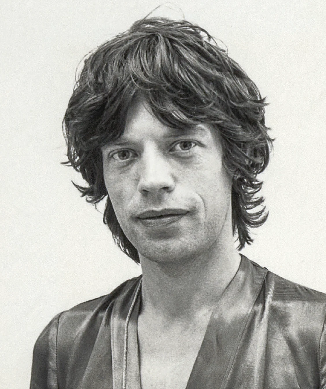 Orta uzunlukta dağınık koyu saçlı bir kişi, parlak açık yakalı bir gömlek giyiyor ve sade bir arka plan önünde kameraya nötr bir ifadeyle bakıyor.