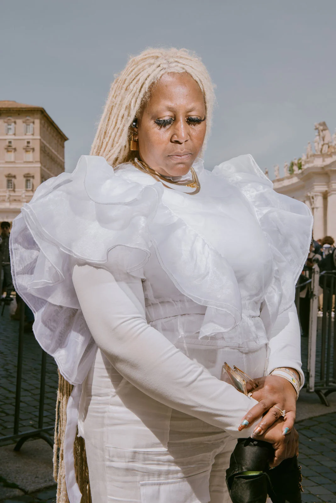 Uzun sarı dreadlock saçlara sahip bir kadın, dramatik beyaz fırfırlı bir elbise içinde güneşli bir günde şehir meydanında duruyor; arka planda tarihi binalar ve mavi gökyüzü yer alıyor.