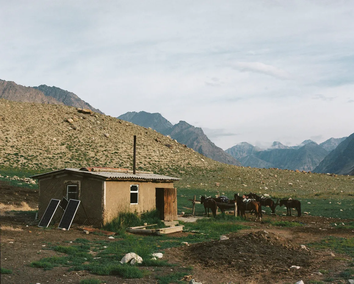Küçük, rustik bir evin dışında iki güneş paneli bulunuyor. Evin çevresinde birkaç at toplanmış. Arka planda kayalık tepeler ve bulutlu gökyüzü altında yükselen dağlar.