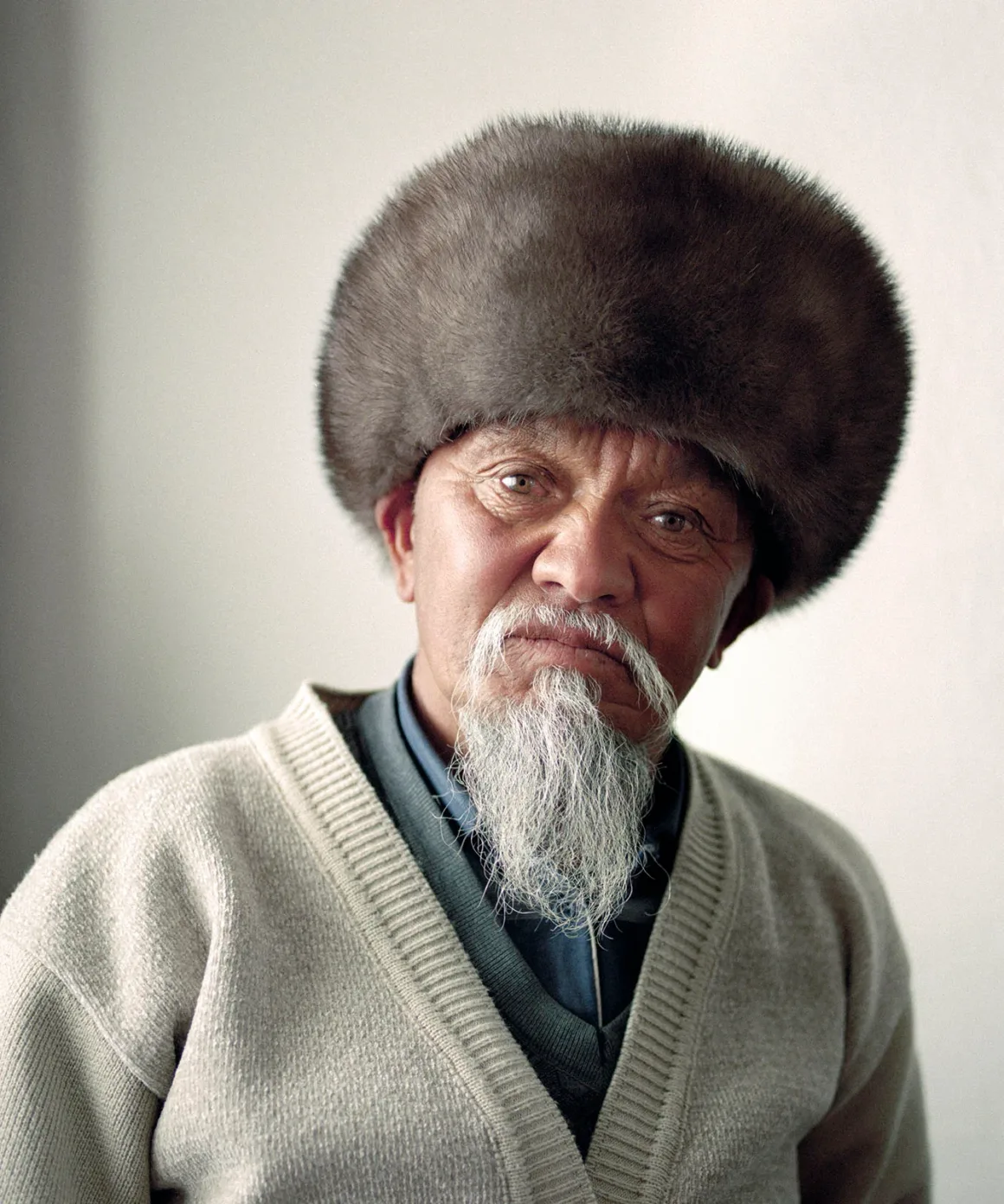 Uzun beyaz sakallı yaşlı bir adam, büyük yuvarlak kürk bir şapka ve yakalı bir gömlek üzerine bej bir hırka giymiş. Düz açık renkli bir arka plan önünde, nötr bir ifadeyle kameraya bakıyor.