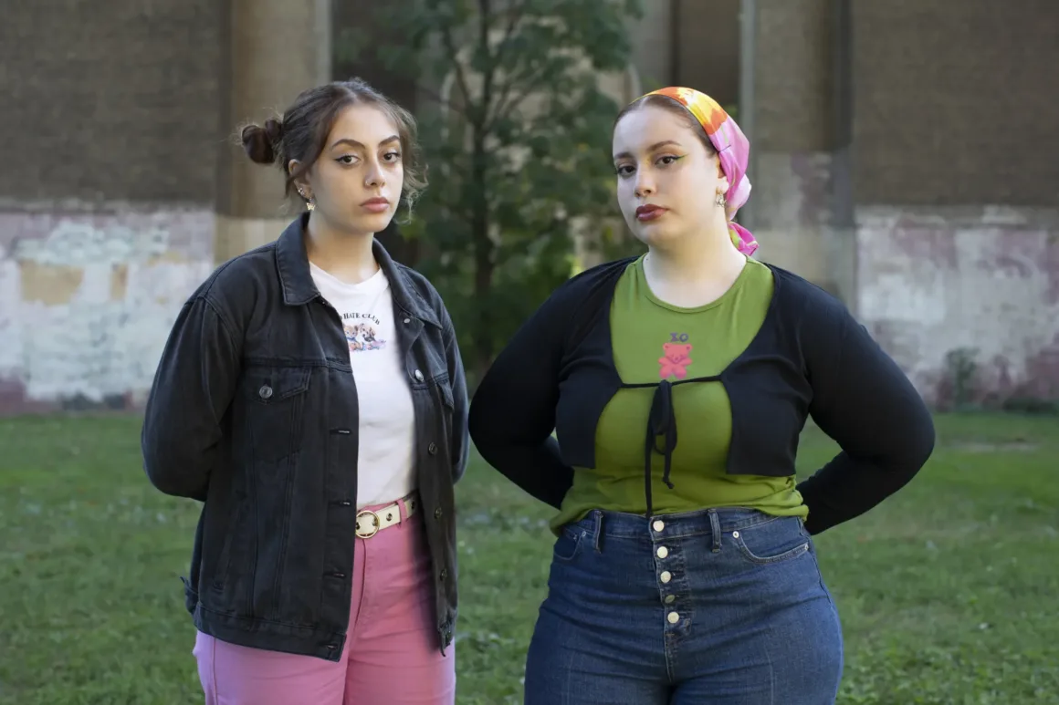 İki genç kadın, çimenlerin üzerinde, grafitilerle kaplı eski bir duvarın önünde duruyor. Biri siyah ceket ve pembe pantolon, diğeri renkli bir eşarp, yeşil üst ve mavi kot pantolon giyiyor. İkisi de kameraya ciddi bir ifadeyle bakıyor.