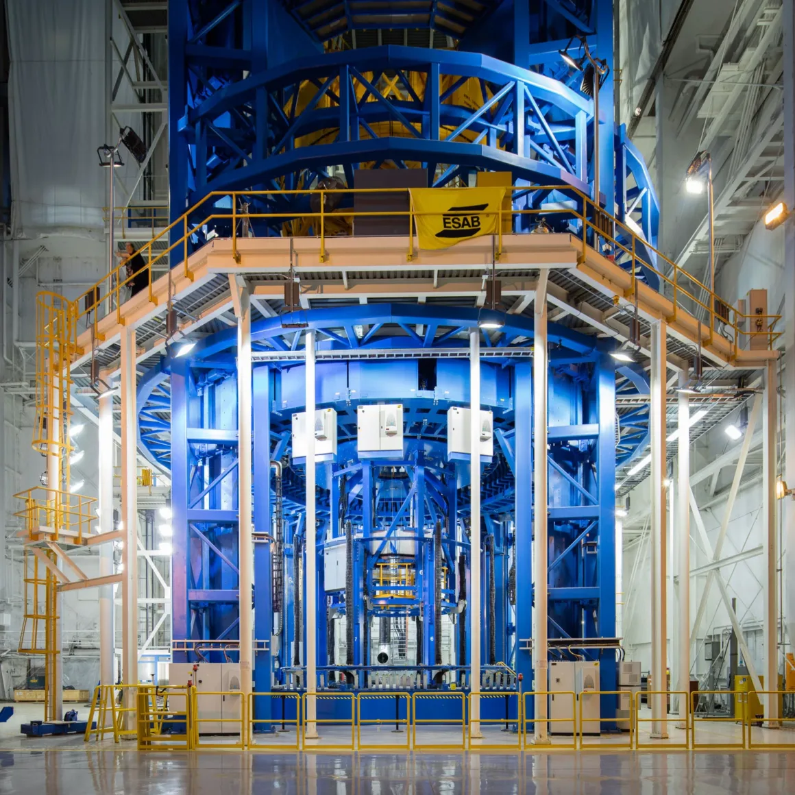 Artemis II’nin Görünmeyen Yüzü: Bir Fotoğrafçı NASA’nın İçini Görüntüledi 8 Parlak ışıkla aydınlatılmış bir tesis içinde, sarı korkuluklar, platformlar ve merdivenlerle çevrili büyük mavi metal bir endüstriyel yapı; etrafta çeşitli borular ve ekipmanlar görülüyor.