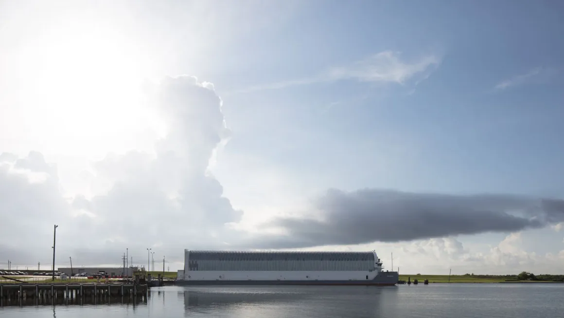 Artemis II’nin Görünmeyen Yüzü: Bir Fotoğrafçı NASA’nın İçini Görüntüledi 2 Büyük, dikdörtgen beyaz bir bina, kıyı yakınında sakin su üzerinde duruyor. Gökyüzünde dramatik bulutlar ve soldan gelen güneş ışığıyla sahne geniş ve huzurlu görünüyor.