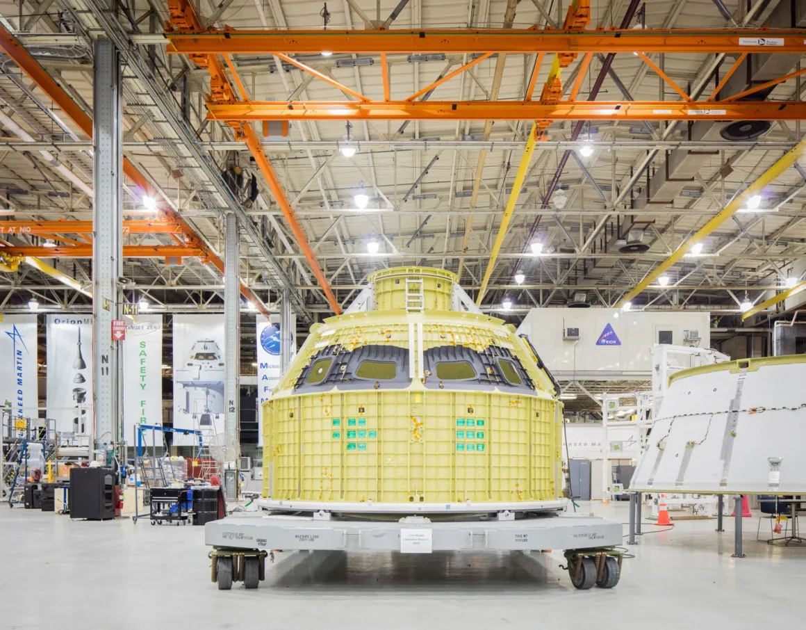 Artemis II’nin Görünmeyen Yüzü: Bir Fotoğrafçı NASA’nın İçini Görüntüledi 10 Sarı dış yüzeye sahip bir uzay aracı modülü, yüksek tavanlı ve aydınlık bir endüstriyel tesis içinde platform üzerinde sergileniyor; arka planda metal kirişler, makineler ve NASA logoları görülüyor.