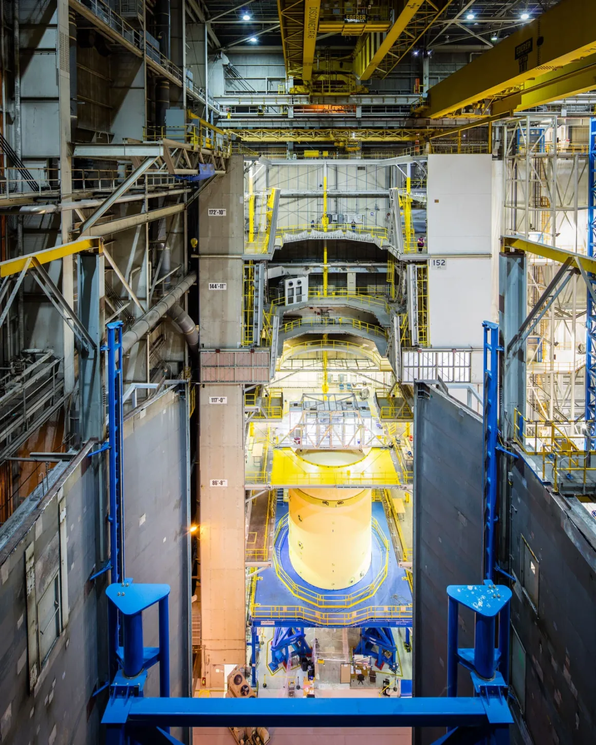 Artemis II’nin Görünmeyen Yüzü: Bir Fotoğrafçı NASA’nın İçini Görüntüledi 3 Yüksek tavanlı geniş bir endüstriyel alanda, sarı metal platformlar, yürüyüş yolları ve korkuluklarla çevrili silindirik bir makine parçası üzerinde çalışan işçiler. Çok katlı yapı ve çeşitli ekipmanlar görülüyor.