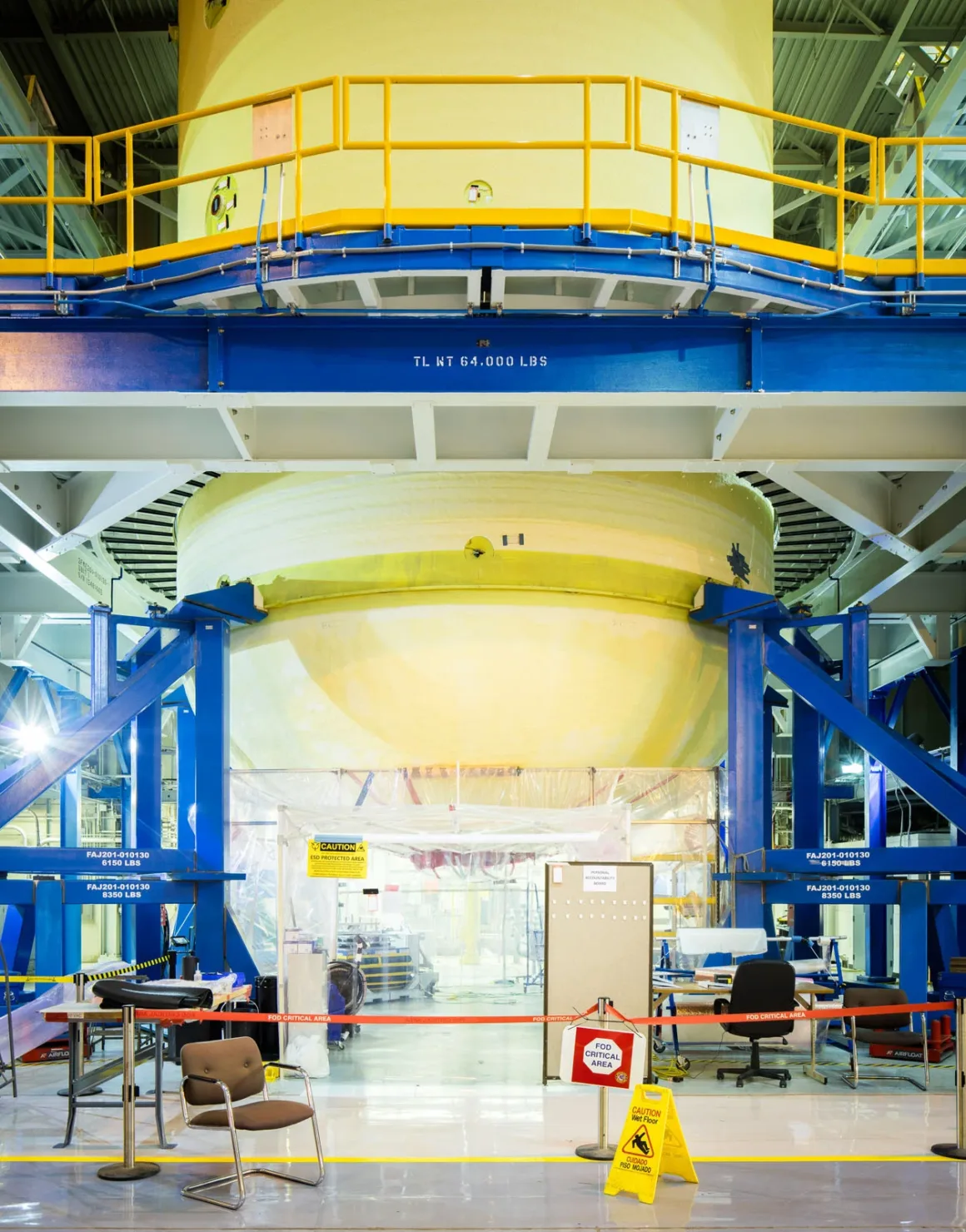 Artemis II’nin Görünmeyen Yüzü: Bir Fotoğrafçı NASA’nın İçini Görüntüledi 4 Büyük sarı silindirik bir roket bileşeni, mavi destek yapılarıyla taşınarak endüstriyel bir tesis içinde havada asılı duruyor. Ön planda sandalyeler, masalar ve bir uyarı tabelası görülüyor.