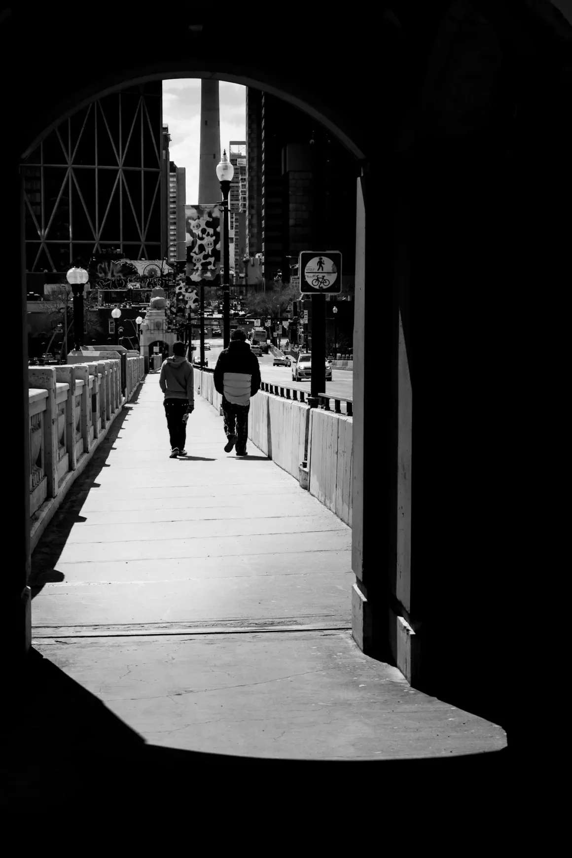 Panasonic Lumix ZS300 İnceleme: Eski Model Daha İyiydi? 32 İki kişi şehir kaldırımında yürüyor. Kadraj, tünel benzeri karanlık bir girişten çerçevelenmiş. Arka planda güneşli bir günde yüksek binalar, sokak lambaları ve yoğun bir cadde görülüyor. Görüntü siyah beyaz.