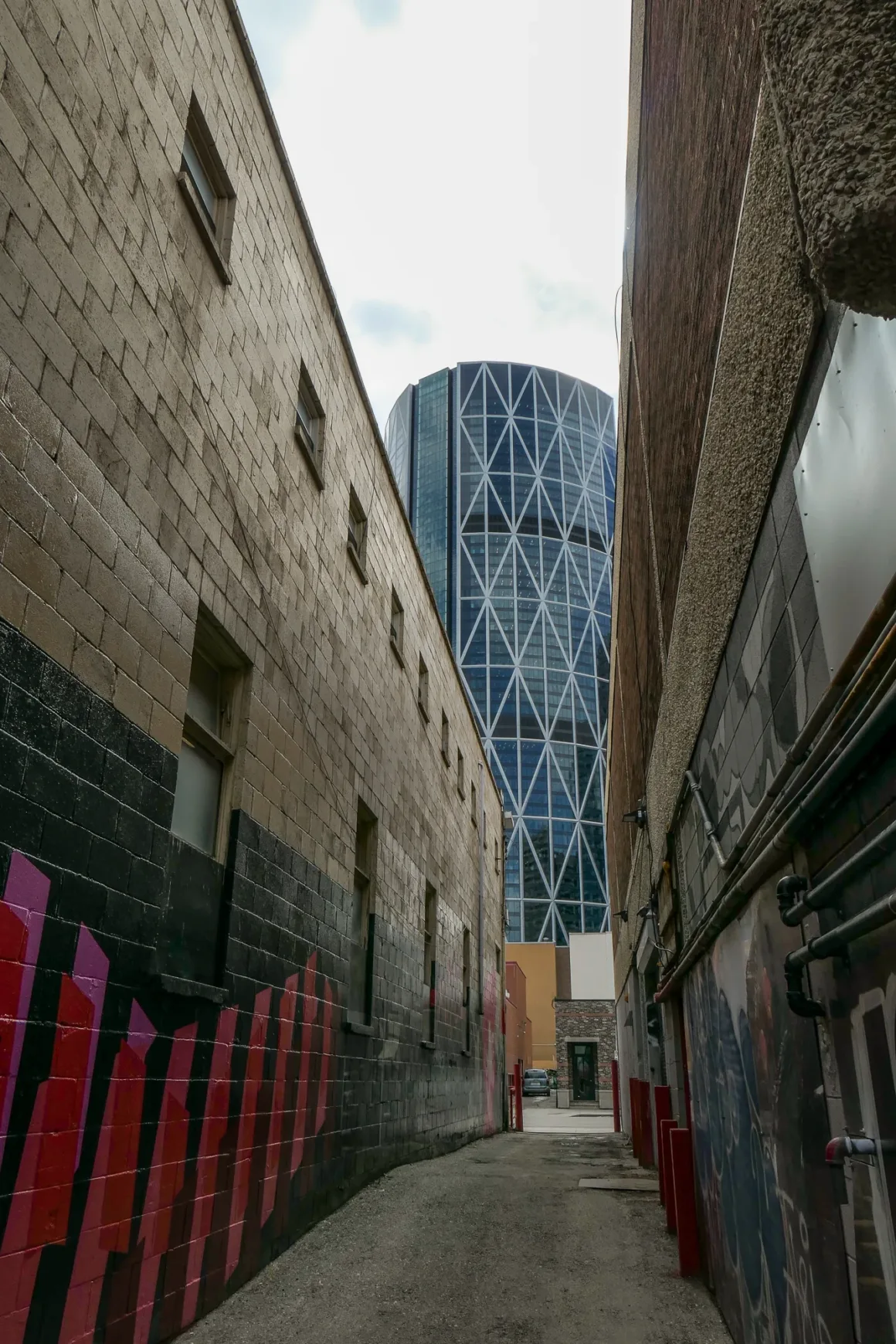 Panasonic Lumix ZS300 İnceleme: Eski Model Daha İyiydi? 26 Bej ve siyah tuğla duvarlarla çevrili dar bir sokak. Duvarlarda grafitiler var ve arka planda bulutlu gökyüzü altında modern bir cam gökdelen görülüyor.