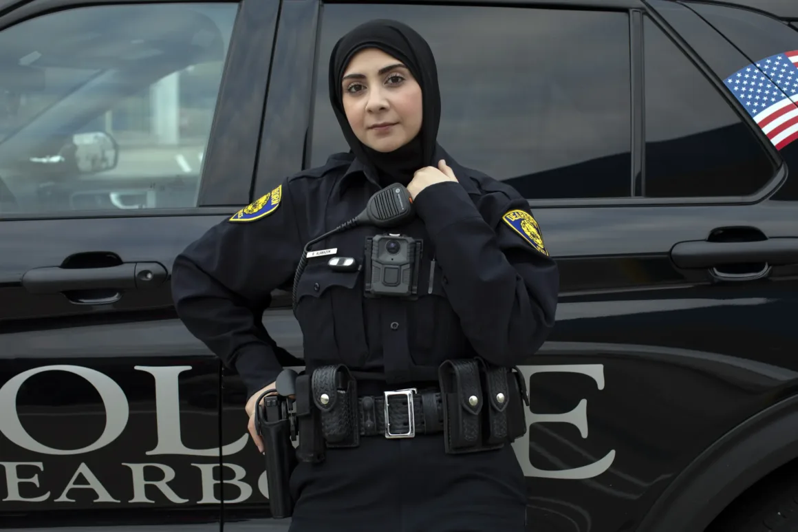 ABD Genelinde 90 Günlük Yolculuk: Bir Fotoğrafçı ‘Müslüman Amerika’yı Belgelemeyi Planlıyor
Siyah üniforma ve başörtüsü takan bir polis memuru, üzerinde Amerikan bayrağı görülen bir polis aracının önünde duruyor. Kameraya kendinden emin bir şekilde bakıyor, bir eli görev kemerinde.