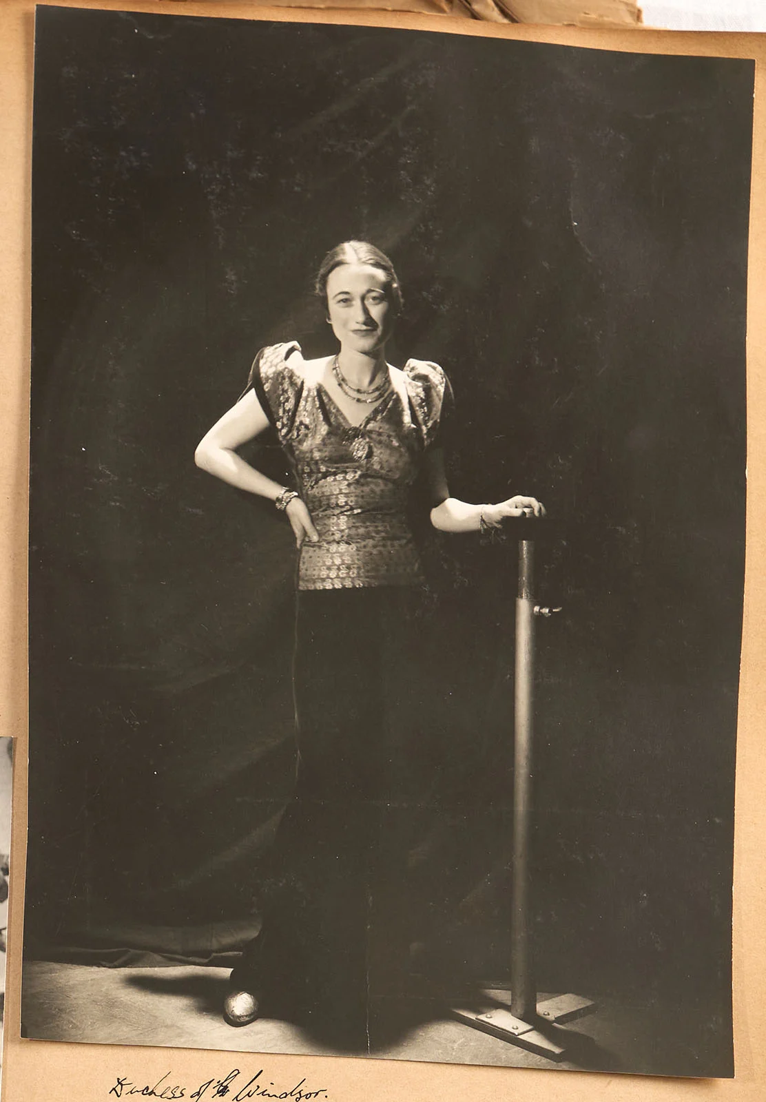 Parlak kısa kollu bir üst ve koyu pantolon giyen bir kadın iç mekânda duruyor; bir eli metal bir standa dayanmış. Arka plan karanlık, kadın kameraya doğru hafif bir gülümsemeyle bakıyor.